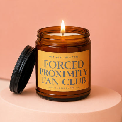 Amber glass candle with lit flame and black lid, labeled Forced Proximity Fan Club.