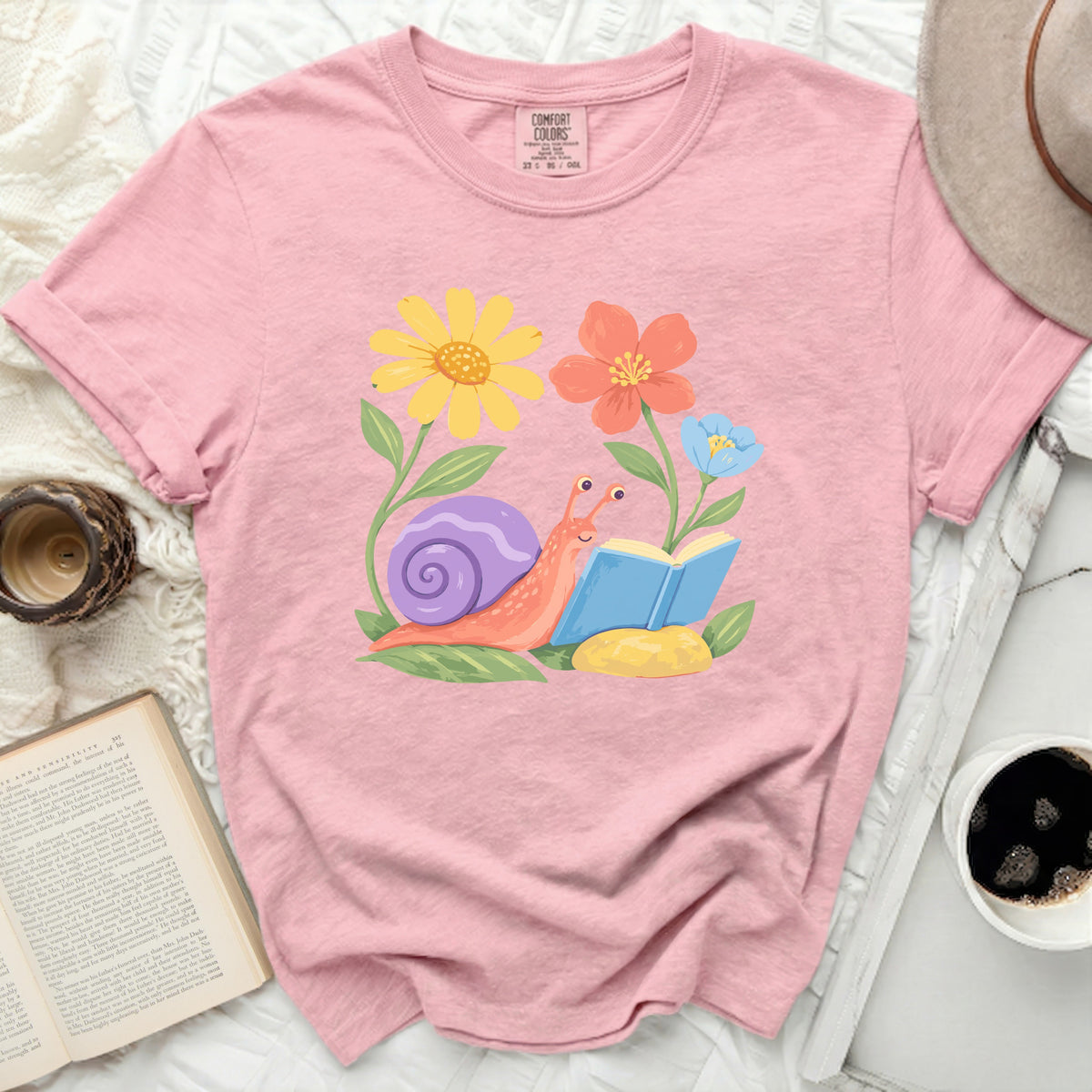 Pink t-shirt with graphic of a snail reading a book among flowers.