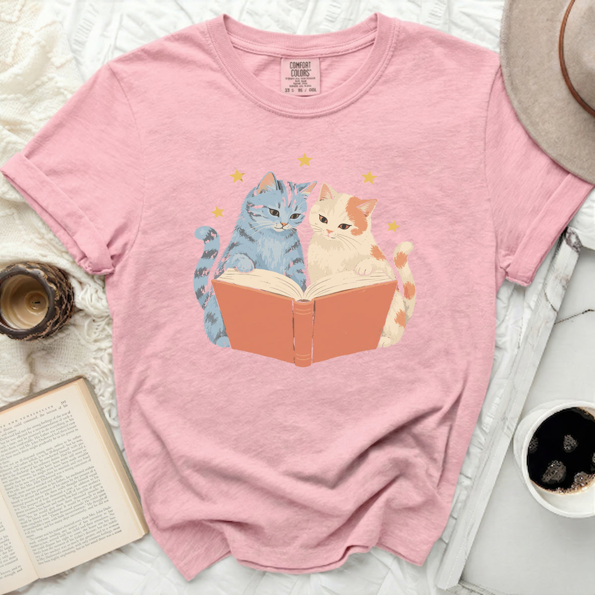 Pink t-shirt featuring a graphic of two cats reading an open book with stars.