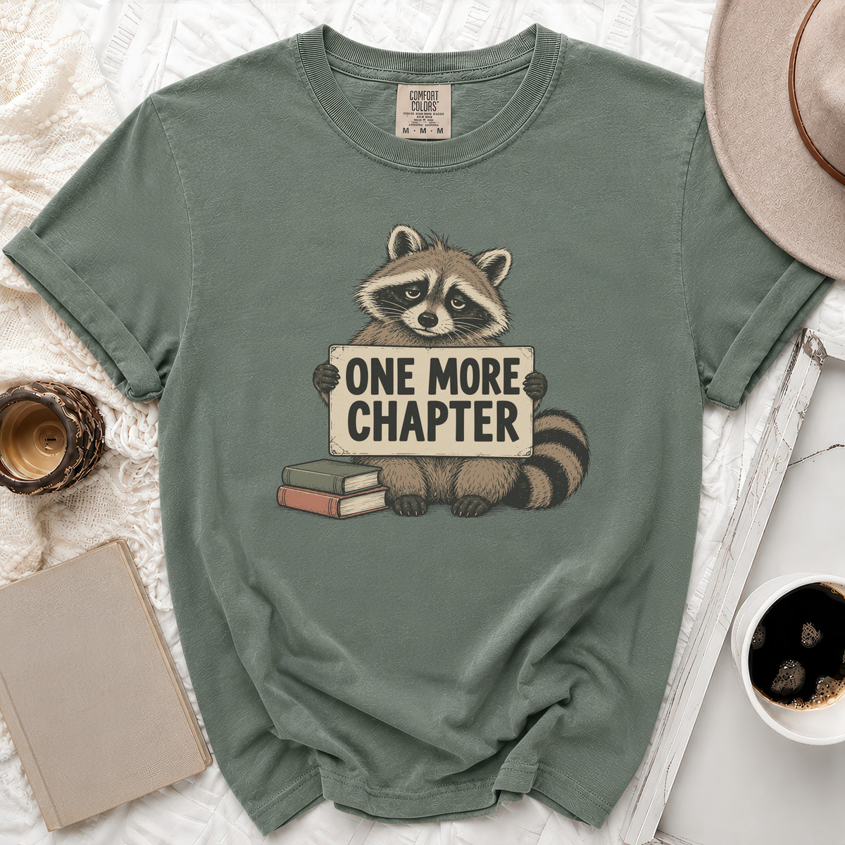 Green t-shirt with a raccoon holding a sign that says "ONE MORE CHAPTER".