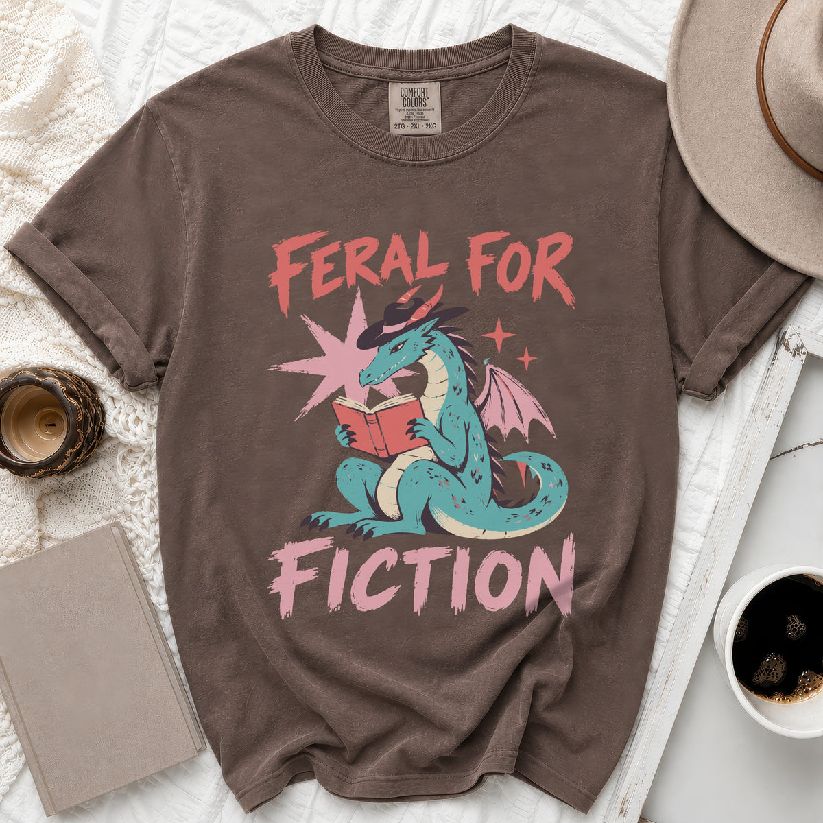 Brown t-shirt with a teal dragon wearing a hat and reading a book, text "Feral for Fiction".