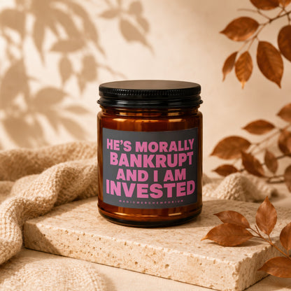 Amber candle with black lid and pink text reading "He's Morally Bankrupt and I Am Invested".