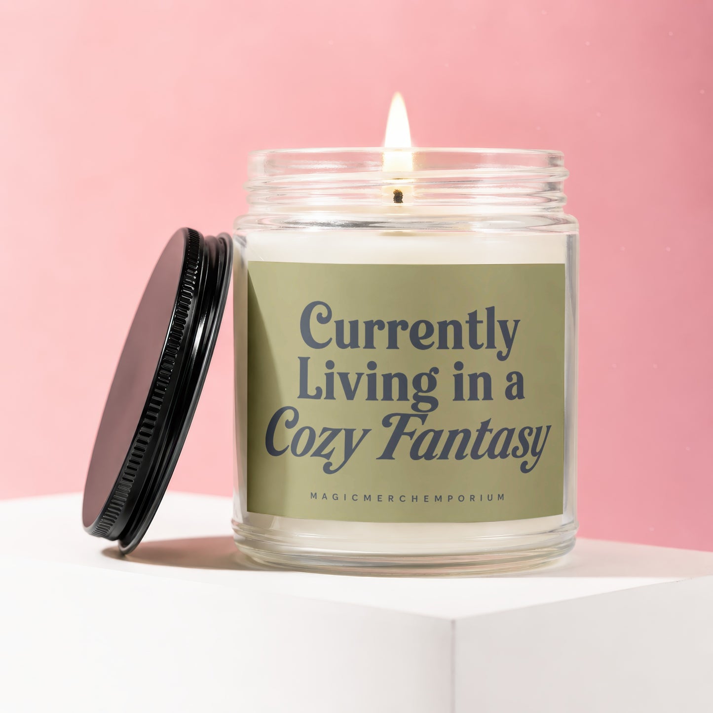 Glass candle with lit wick and black lid, labeled "Currently Living in a Cozy Fantasy".