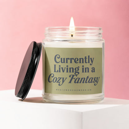 Glass candle with lit wick and black lid, labeled "Currently Living in a Cozy Fantasy".