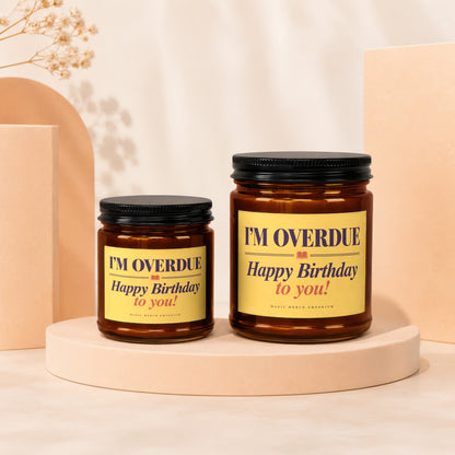 Two amber jar candles with black lids on peach pedestals, one larger than the other, featuring yellow labels that read "I'M OVERDUE Happy Birthday to you!".
