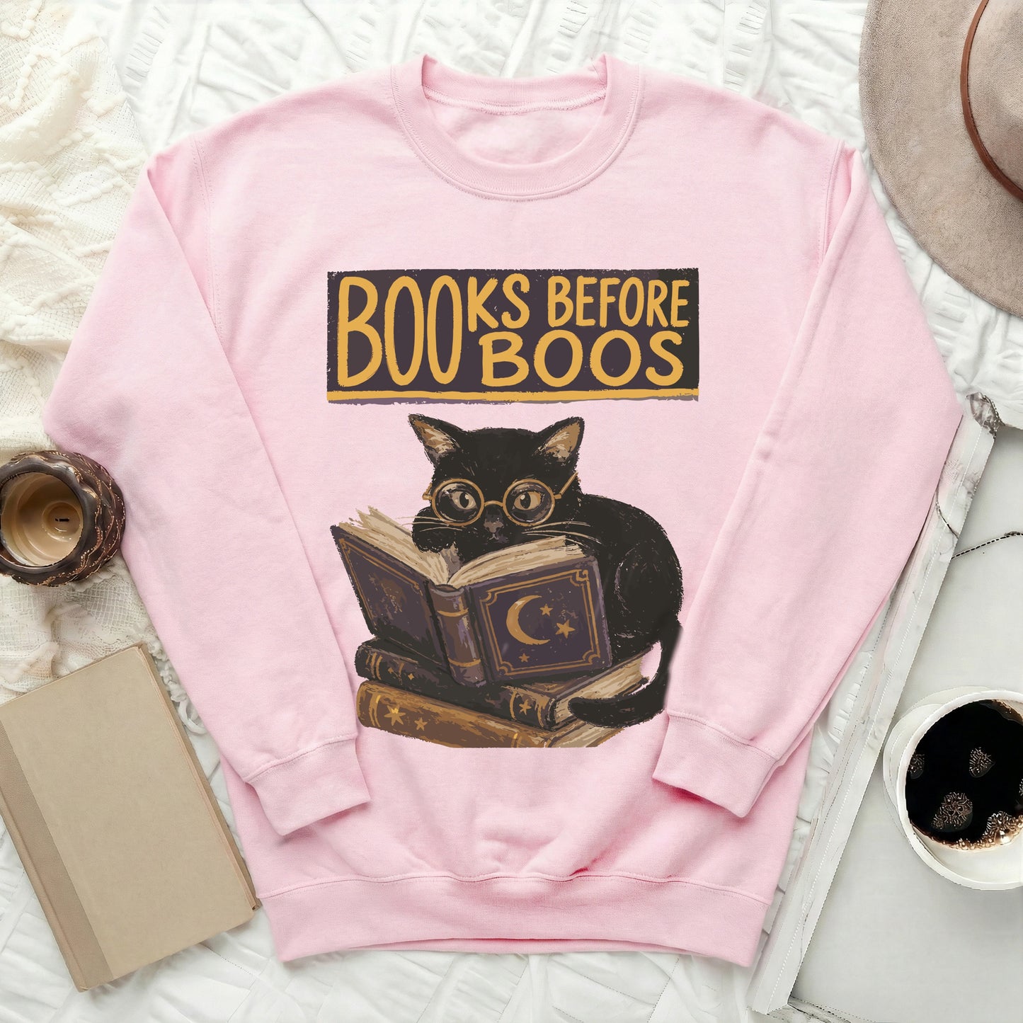 Pink sweatshirt with a graphic of a black cat reading books, text "Books Before Boos".