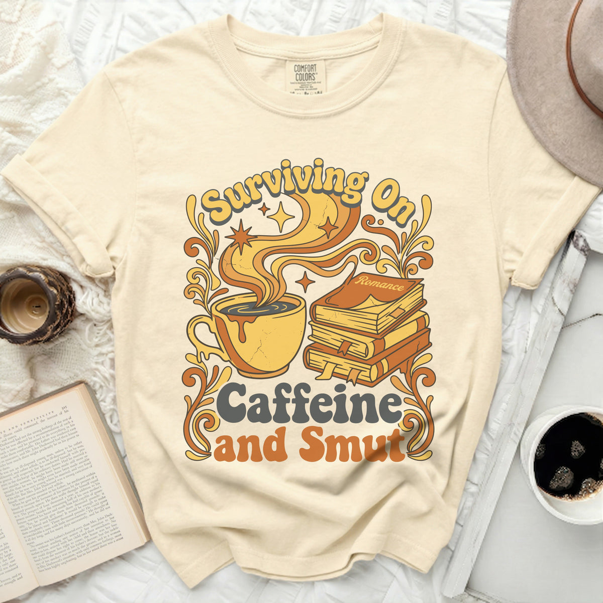 Cream t-shirt with a graphic of a coffee cup, books, and the text "Surviving On Caffeine and Smut".
