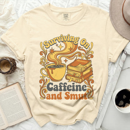 Cream t-shirt with a graphic of a coffee cup, books, and the text "Surviving On Caffeine and Smut".