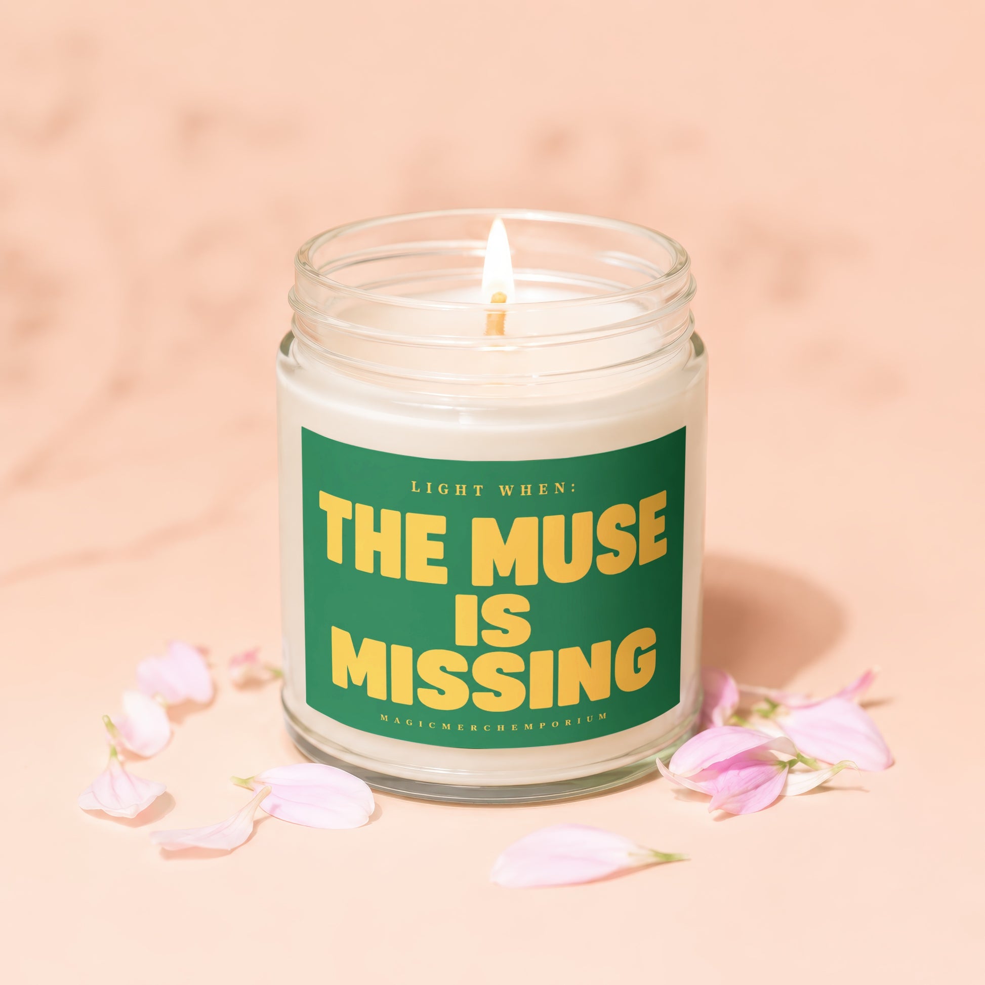 Glass jar candle with lit wick, green label reading "Light When: The Muse Is Missing Magic Merch Emporium."