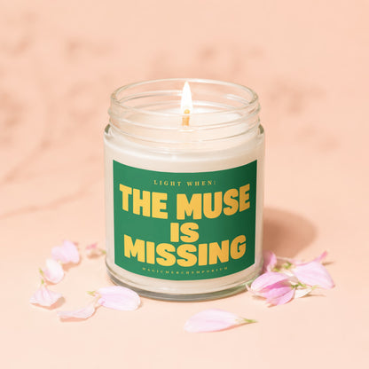 Glass jar candle with lit wick, green label reading "Light When: The Muse Is Missing Magic Merch Emporium."