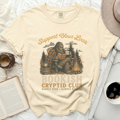 Bigfoot reading a book and drinking from a mug with "Support Your Local Bookish Cryptid Club" printed on the shirt.