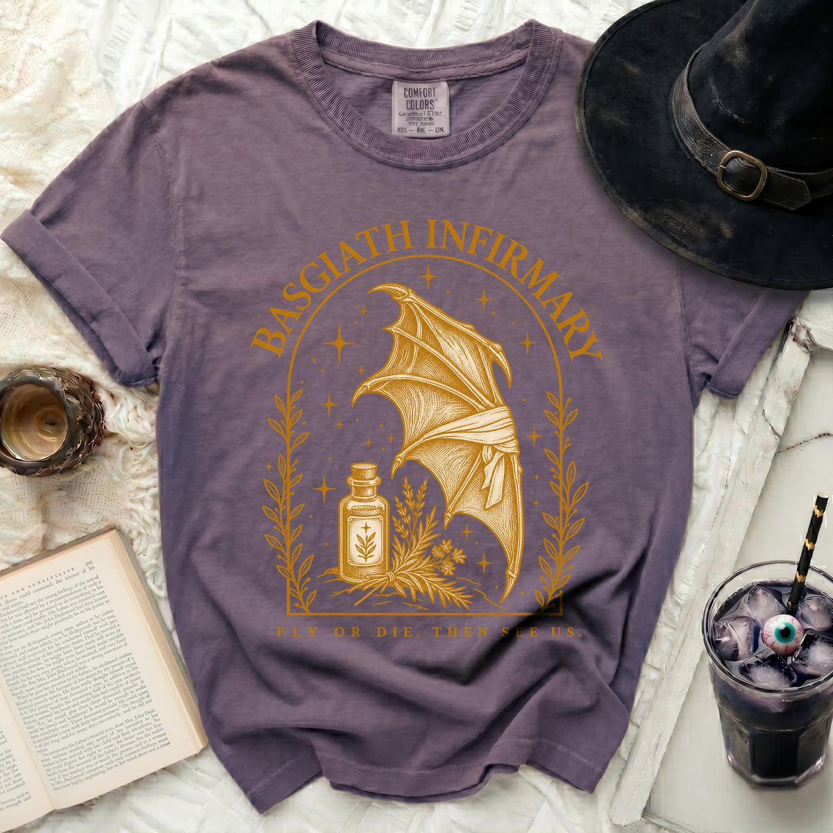 A purple t-shirt featuring a gold graphic of a dragon wing, potion bottle, and Basgiath Infirmary text.