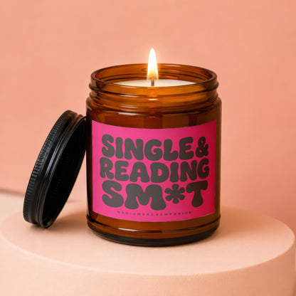 Amber jar candle with lit flame and a pink label reading "SINGLE & READING SM*T".