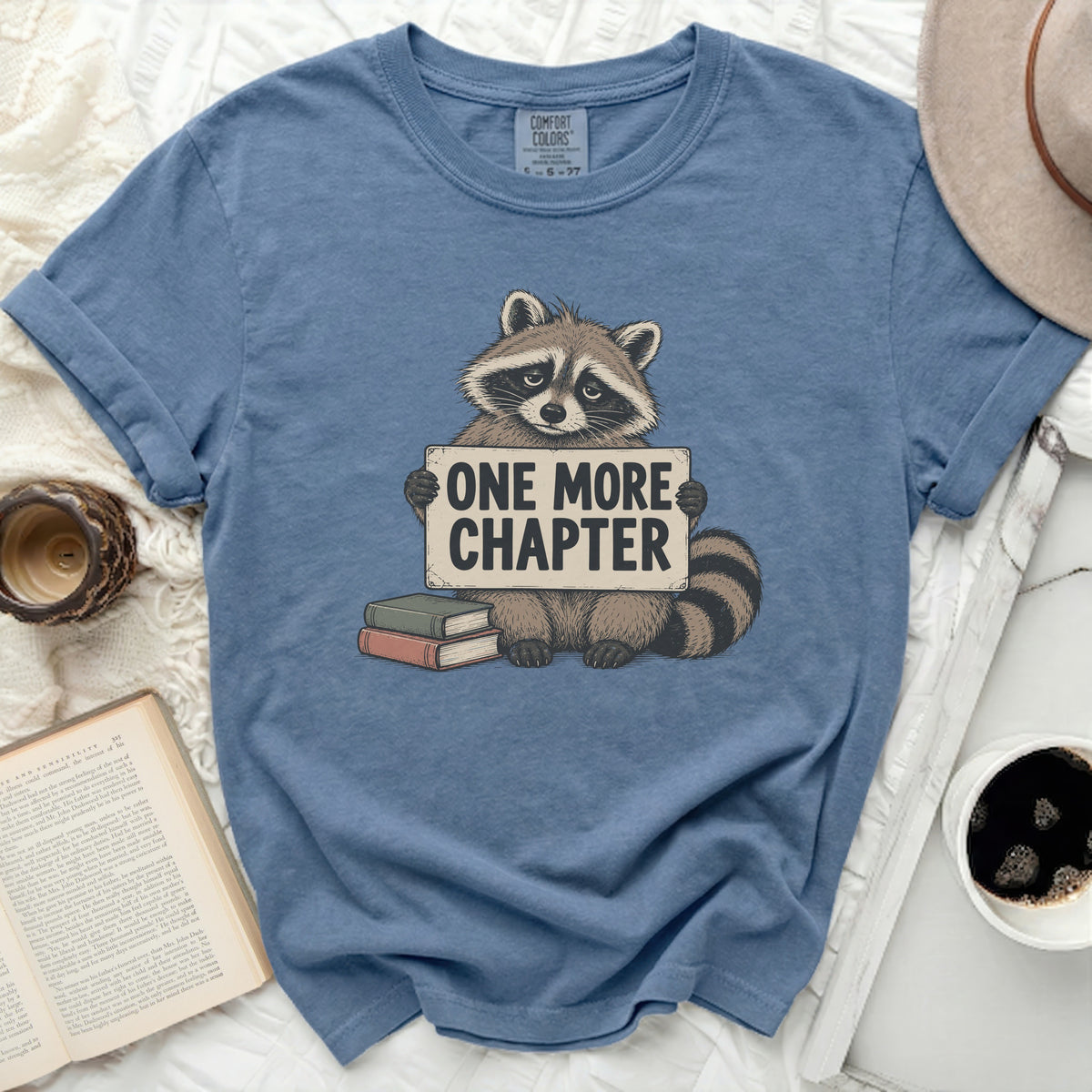 Blue t-shirt with raccoon graphic holding "ONE MORE CHAPTER" sign and books.