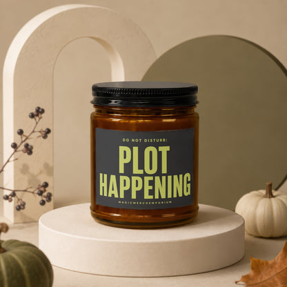 Amber jar candle with "PLOT HAPPENING" text on label, displayed with decorative pumpkins.