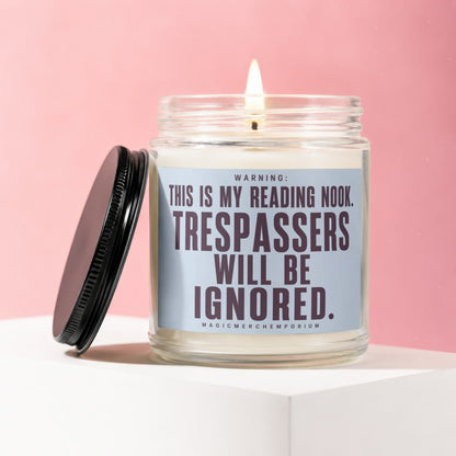 Glass candle with lit wick and black lid featuring text "WARNING: THIS IS MY READING NOOK. TRESPASSERS WILL BE IGNORED."