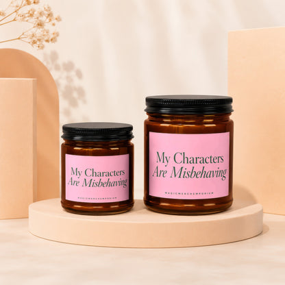 Two amber glass candles with pink labels reading "My Characters Are Misbehaving".