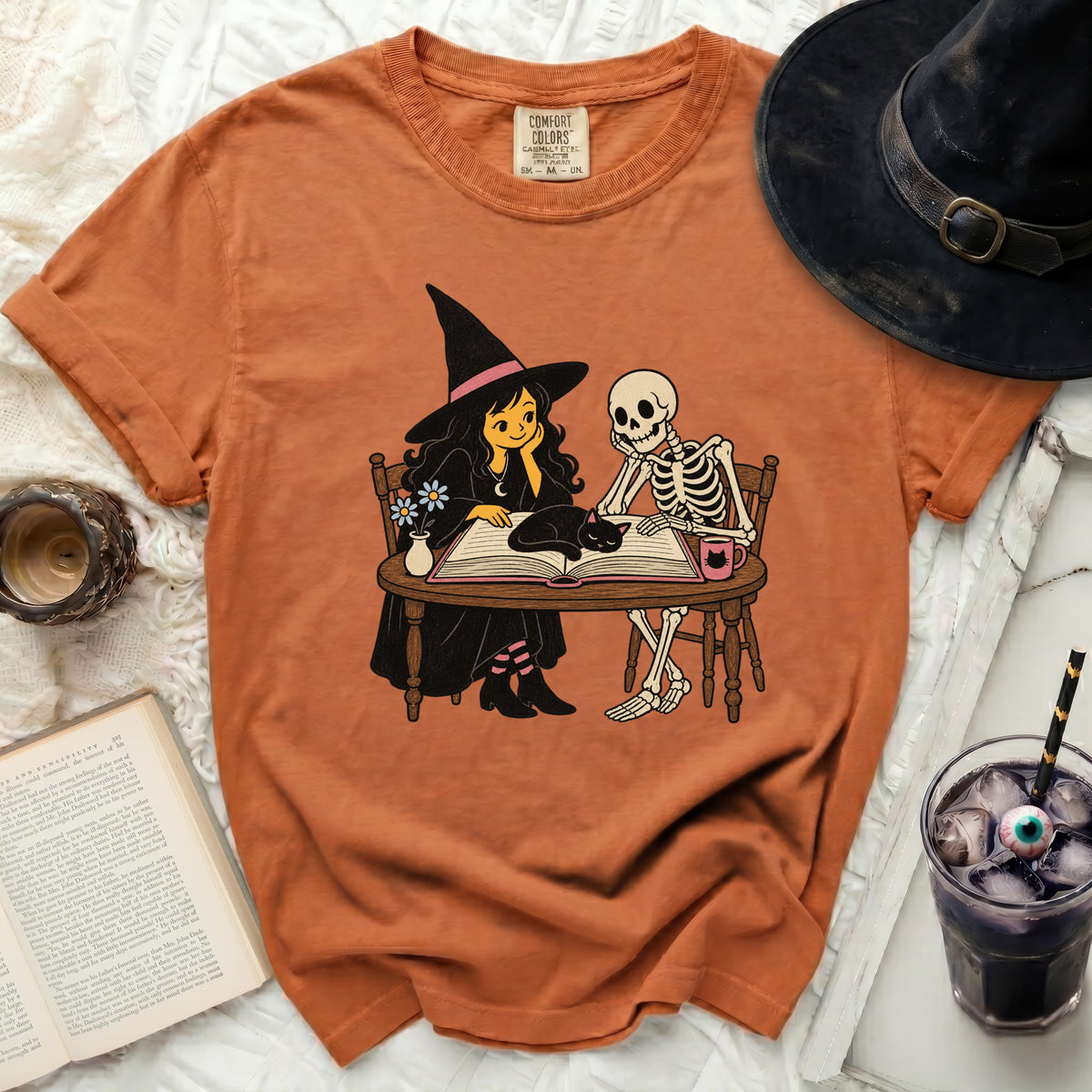 Orange t-shirt featuring a witch, skeleton, and cat reading a book.
