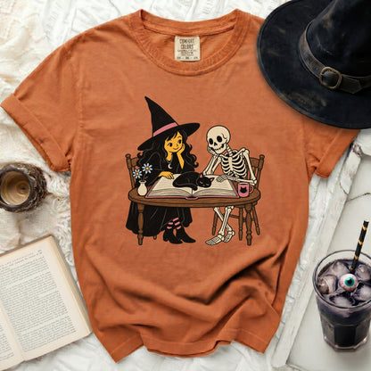 Orange t-shirt featuring a witch, skeleton, and cat reading a book.
