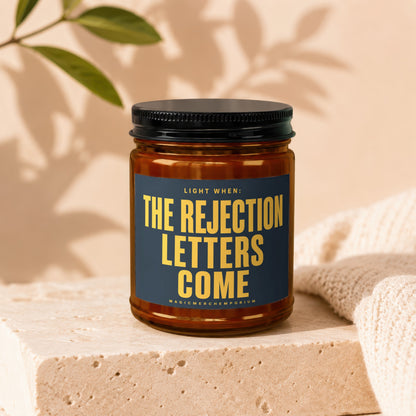 Amber glass candle with black lid, featuring a dark blue label that reads: "LIGHT WHEN: THE REJECTION LETTERS COME"