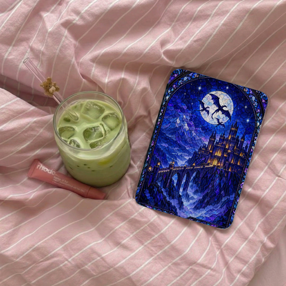 A Kindle case featuring a blue stained-glass castle design lies on a pink striped bedspread next to a glass of iced matcha and a pink lip balm.