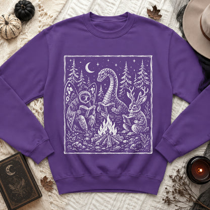 Purple sweatshirt with white graphic of mythical creatures reading by campfire at night.