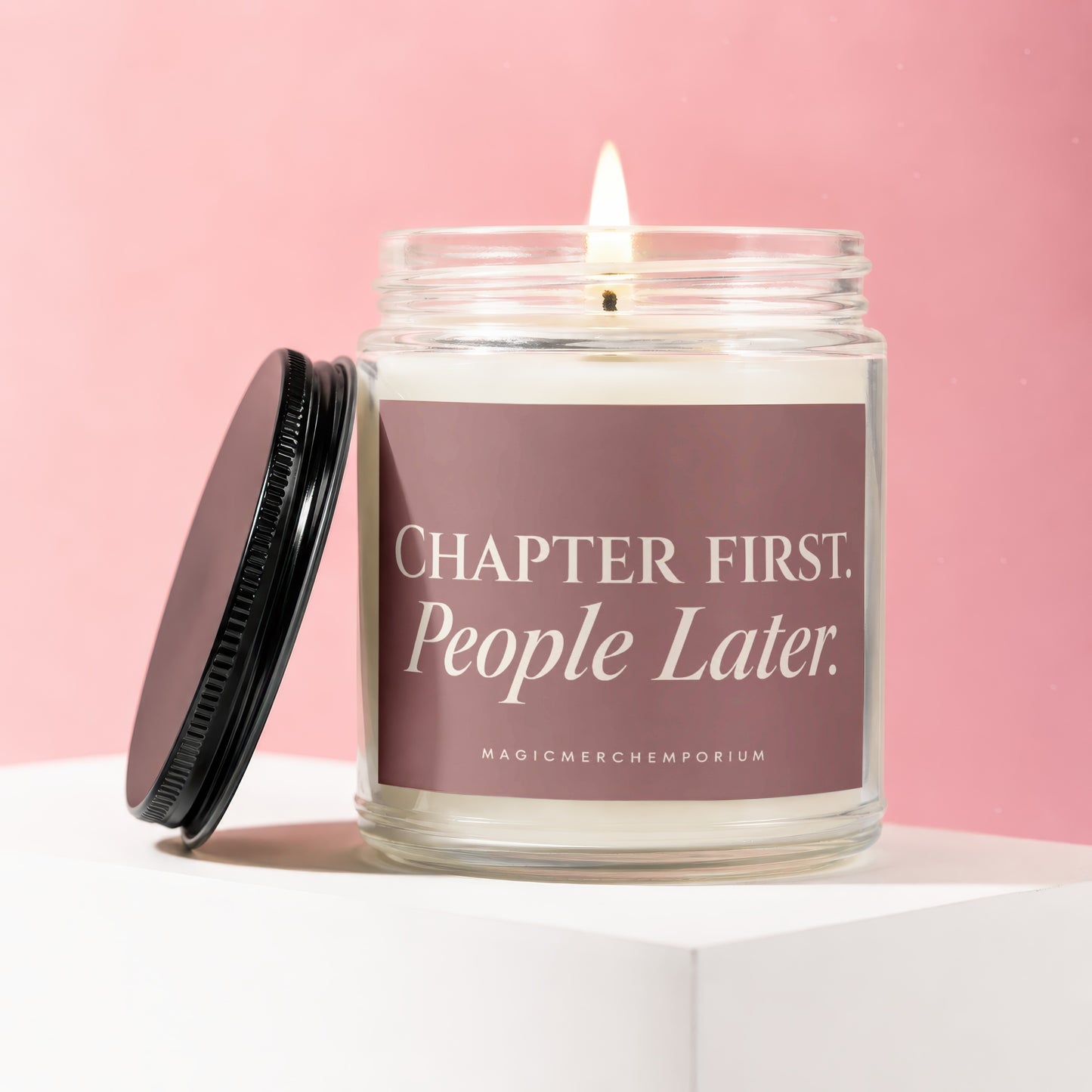 Glass candle with lit wick, black lid, and label that reads "Chapter First. People Later. MAGICMERCHTEMPORIUM."