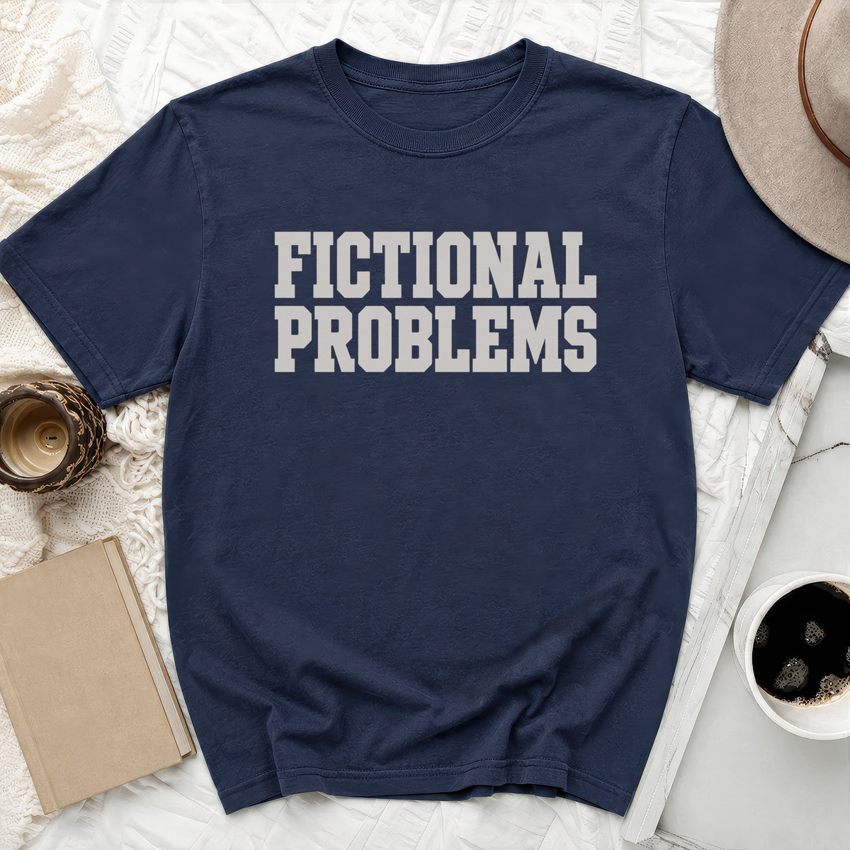 A navy blue short-sleeve t-shirt with block lettering reading FICTIONAL PROBLEMS laid flat on a white textured surface with a book, candle, hat, and coffee cup.