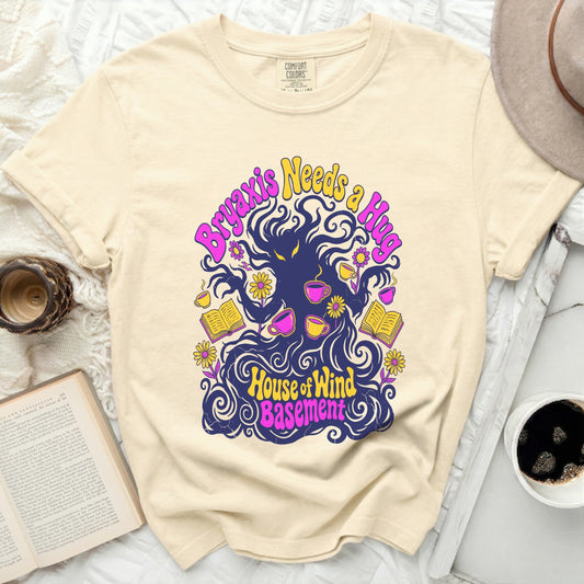 Cream t-shirt with a graphic featuring dark wavy hair, yellow flowers, books, and cups. Text reads "Bryaxis Needs a Hug" and "House of Wind Basement."