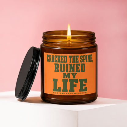 Amber candle with lit wick and black lid, reading "CRACKED THE SPINE, RUINED MY LIFE".