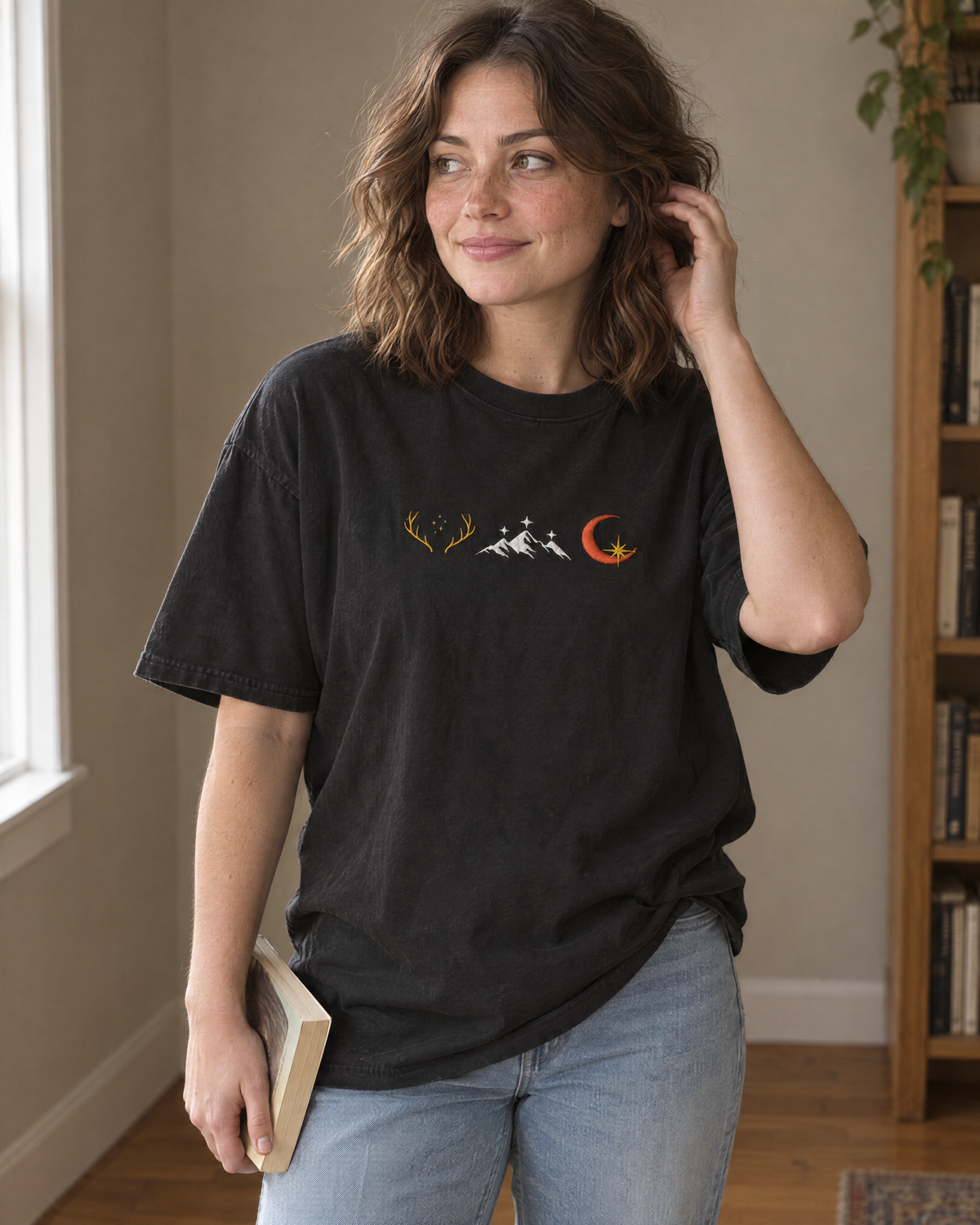 Woman wearing the black Comfort Colors tee with an embroidered row of three emblems (golden antlers with stars, white mountains with stars, crescent moon with a gold star) on the chest