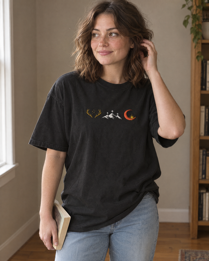 Woman wearing the black Comfort Colors tee with an embroidered row of three emblems (golden antlers with stars, white mountains with stars, crescent moon with a gold star) on the chest