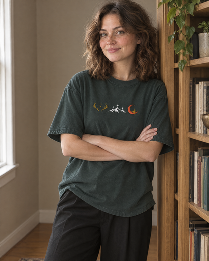 Woman leaning on a bookshelf in the Blue Spruce green tee, embroidered row of three emblems (golden antlers with stars, white mountains with stars, crescent moon with a gold star) on the chest