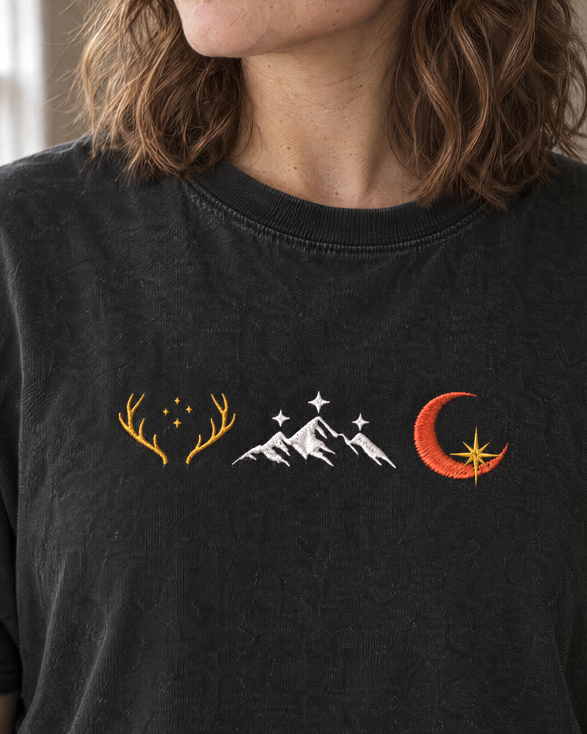 Close-up of the stitched embroidered row of three emblems (golden antlers with stars, white mountains with stars, crescent moon with a gold star) on the chest on a black tee