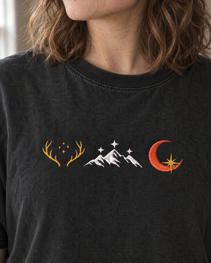 Close-up of the stitched embroidered row of three emblems (golden antlers with stars, white mountains with stars, crescent moon with a gold star) on the chest on a black tee