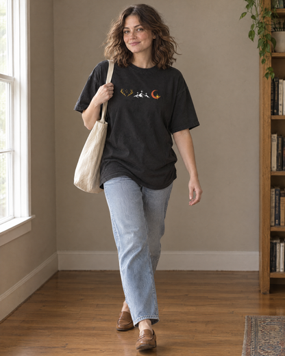 Woman walking in the Pepper charcoal tee, embroidered row of three emblems (golden antlers with stars, white mountains with stars, crescent moon with a gold star) on the chest